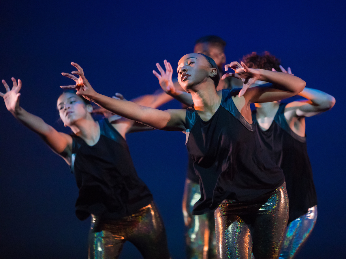 Four dancers of color wearing black sleeveless tops and metallic pants are in front of a blue background. Their bodies are twisted and angled to the left. Their hands are clawed with one arm reaching forward and the elbow of the other arm pulled back.