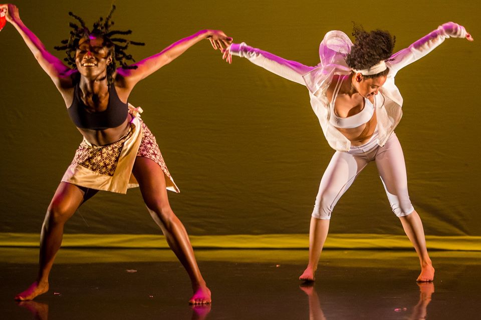 Two women of color dance in front of a golden backdrop. They stand with their legs apart and sitting into one hip. Their torsos are pitched forward and their arms are spread wide. The woman on the left has her face lifted towards the audience. The woman on the right has her face turned to the right. Christine Cano is the woman on the right. 